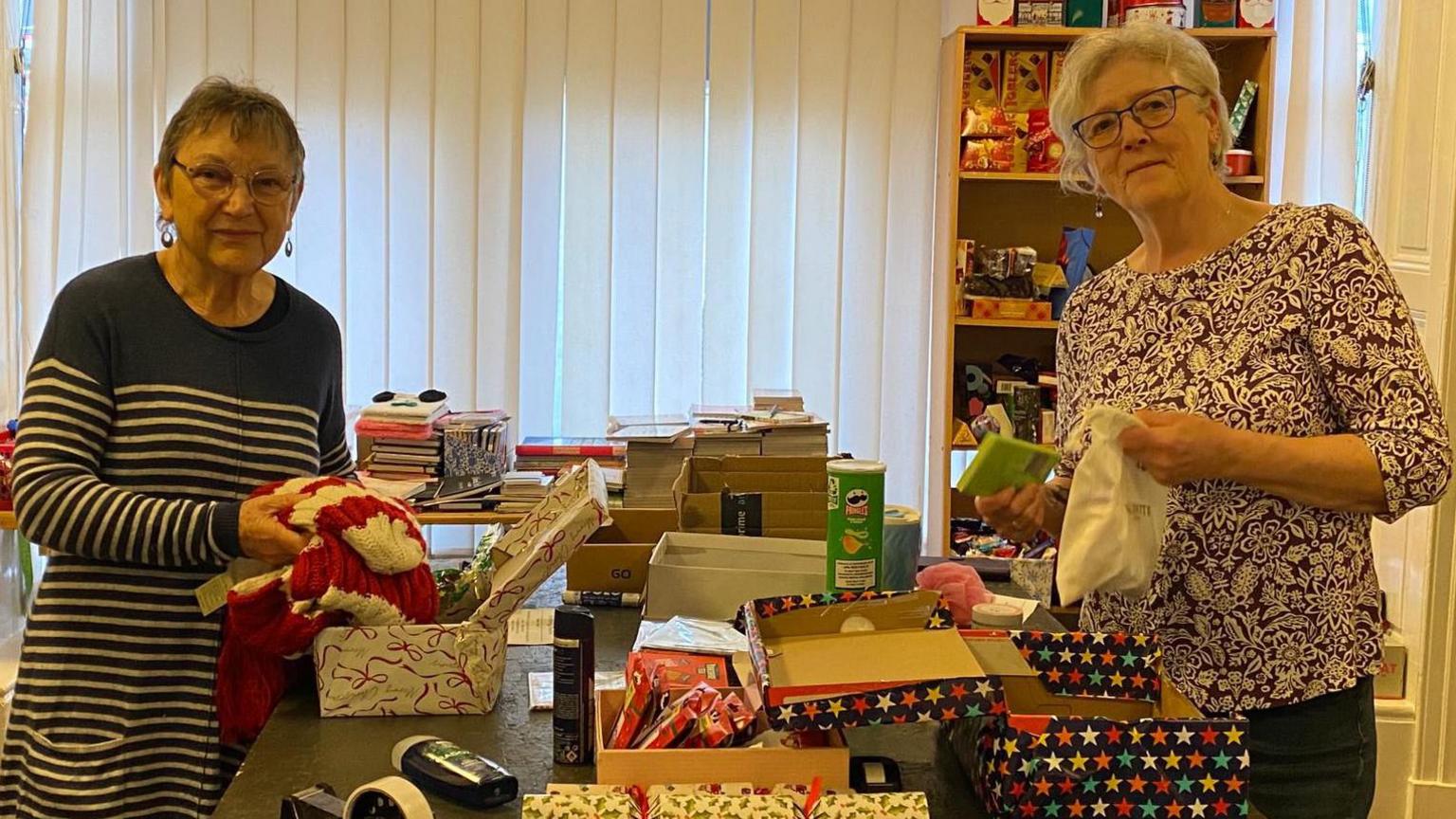Two senior women, volunteers for Sheffield Shoebox Appeal, are stood either side of a table with shoeboxes, wrapping paper and gift items to fill the boxes.
