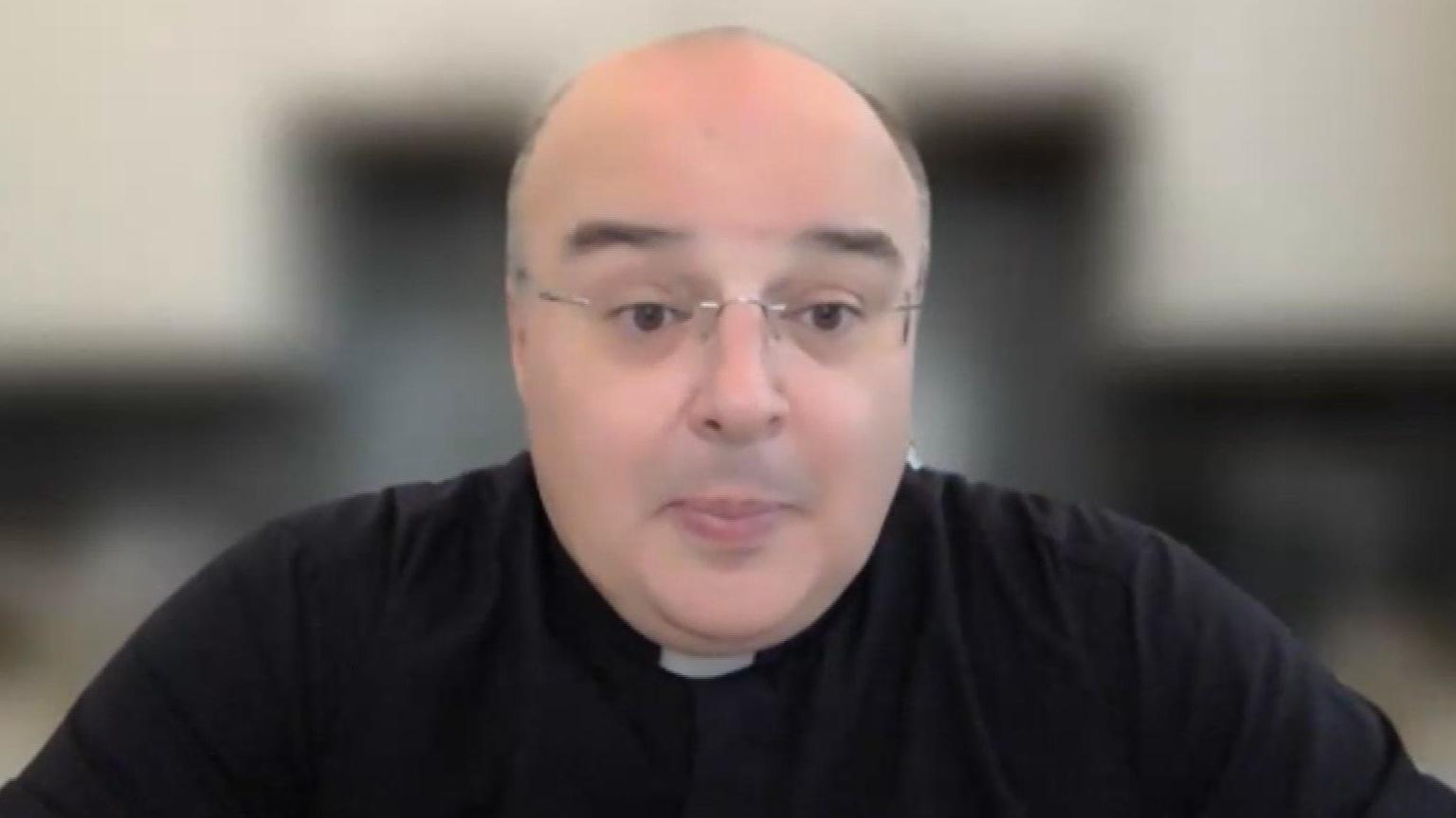 Fr Domenico is speaking on a Teams call. He wears glasses and a priests' clothing. The background behind him is blurred.