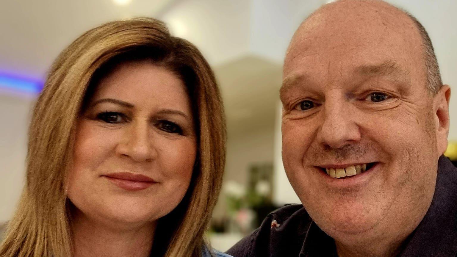 Pam and Kevin smile at the camera. Pam has long brown hair and is wearing pink lipstick, while Kevin has short grey hair and is clean shaven. The background is blurred behind them.