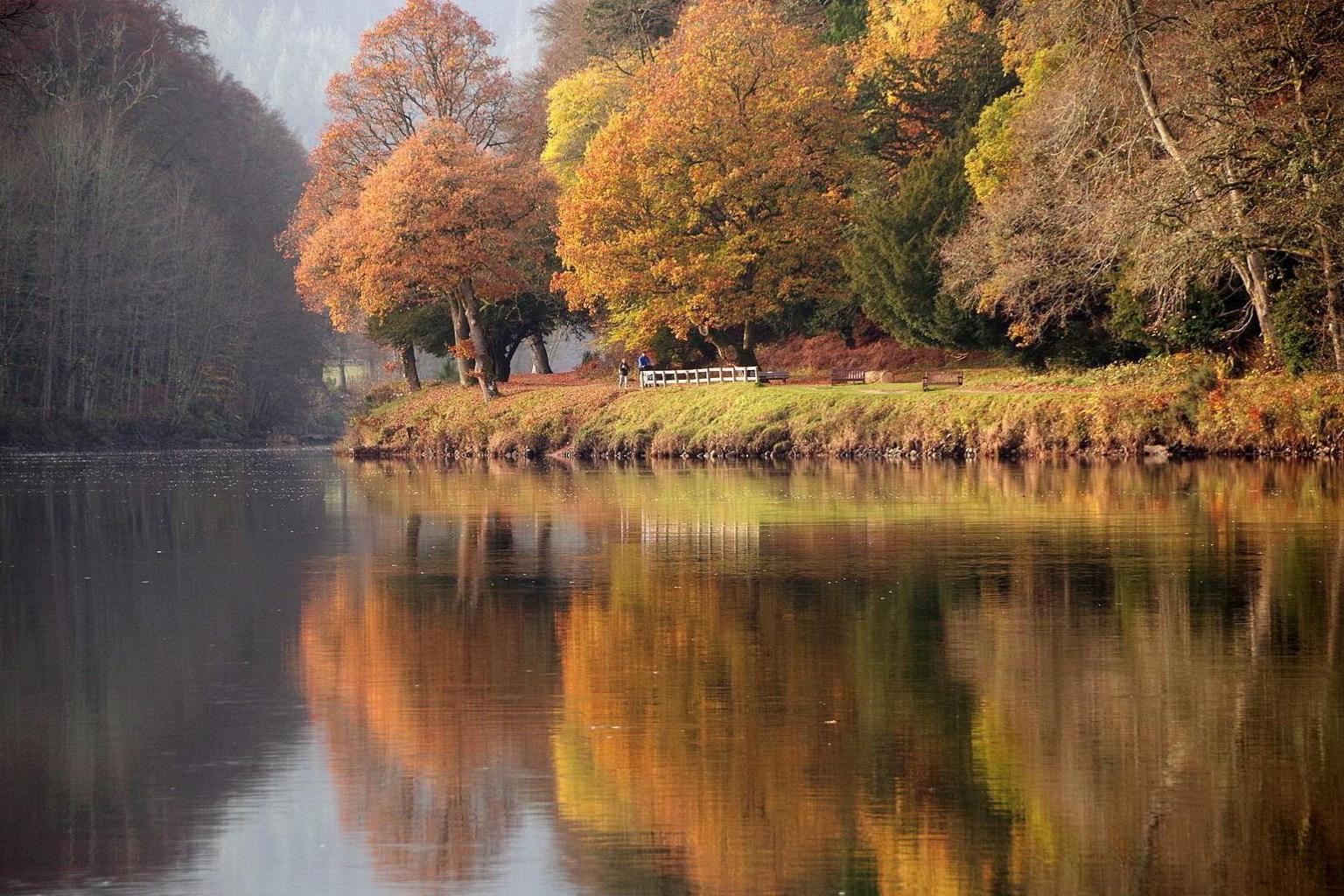 Trees with leaves of orange, red and gold are reflected in water below. There are two people walking near a wooden fence.