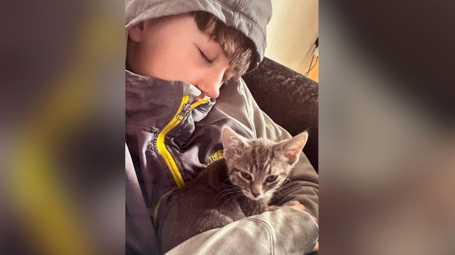 Christopher McBurnie is photographed with his eyes closed with his hood up over his head. A small grey tabby cat is sitting in his arms.