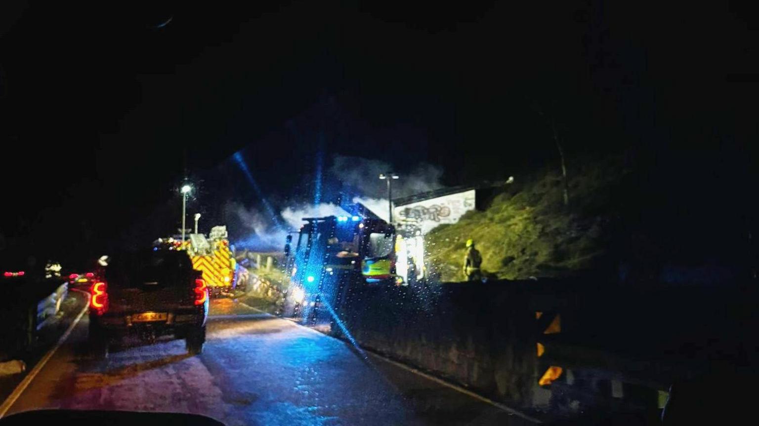 Emergency service response vehicles parked roadside with a graffitied building in the distance and smoke coming from it