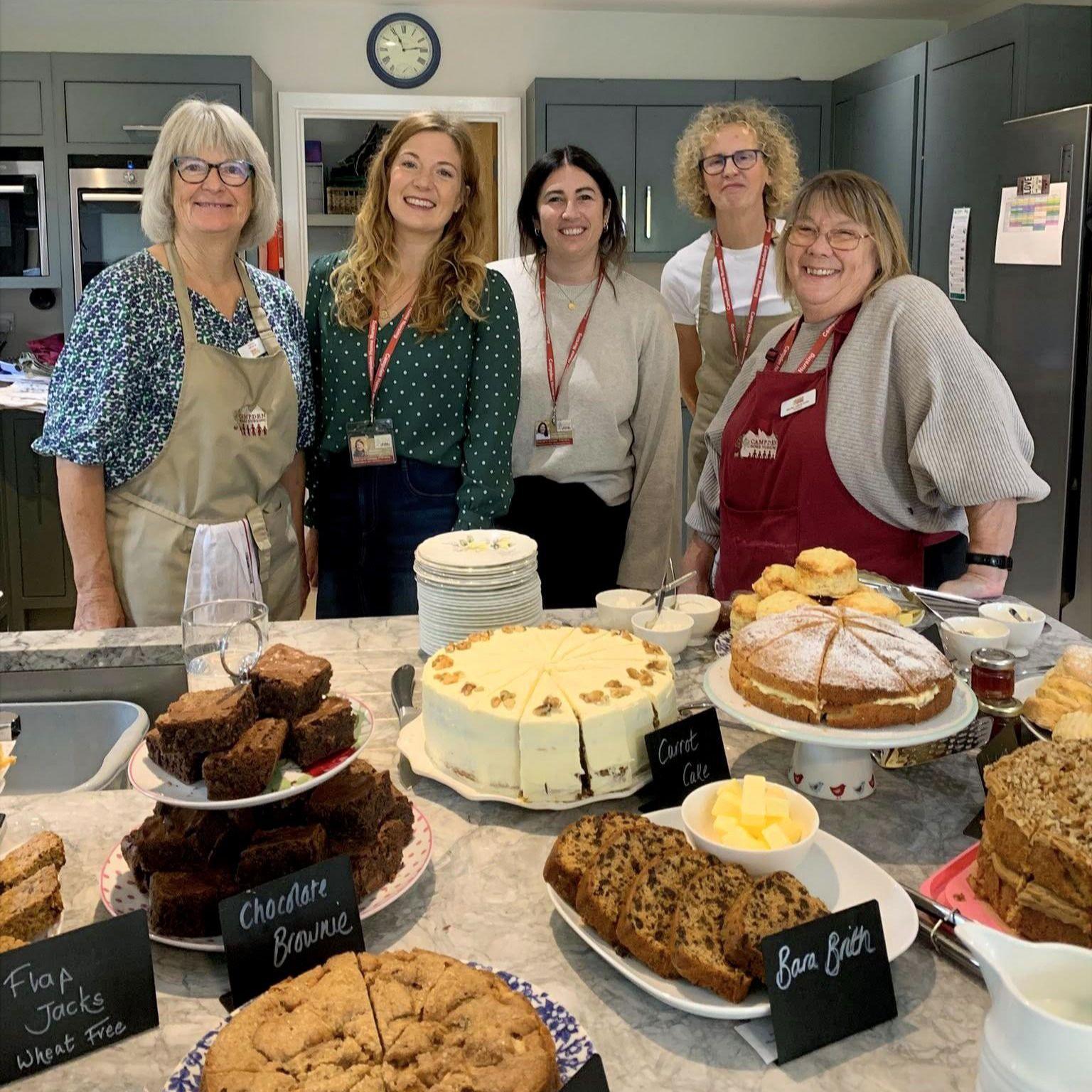 Five women standing in front of a variety of homemade cakes and brownies. They are all smiling.