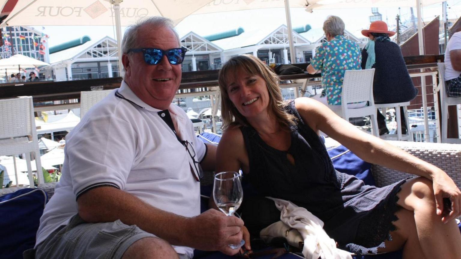 Geoff Pike, wearing sunglasses and a white polo shirt, sat on a sofa next to wife Helen who is in a dark dress.