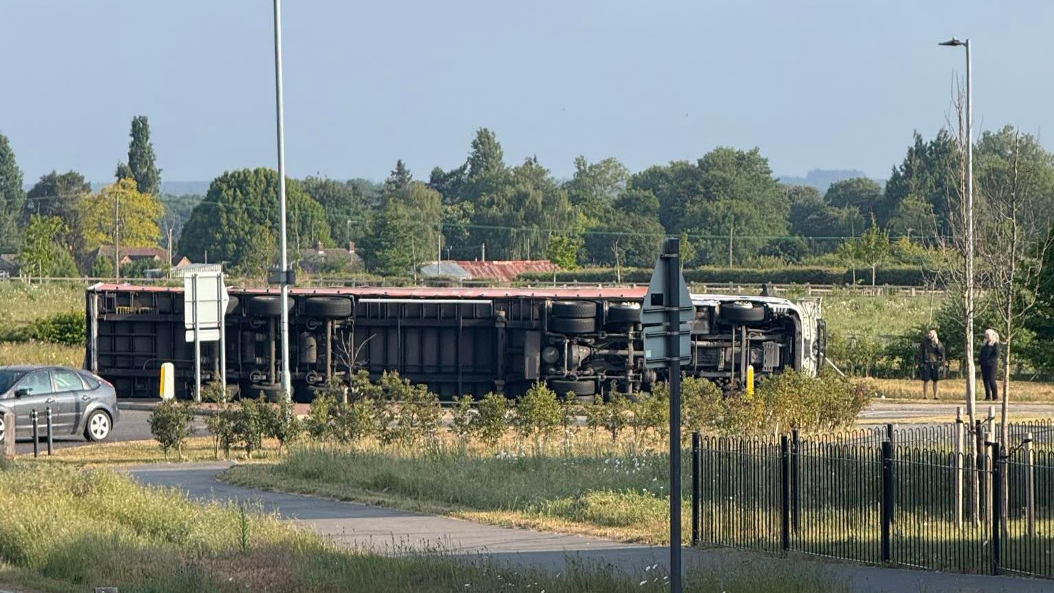 An overturned lorry rests on the side of a road. Its undercarriage can be seen. Another car drives nearby as people stand on the side of the road and watch on.