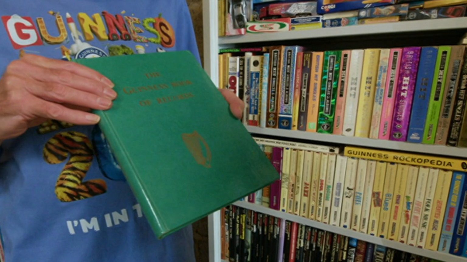 A man is holding a green Guinness World Record book into a camera next to a book shelf filled with more Guinness World Record books.