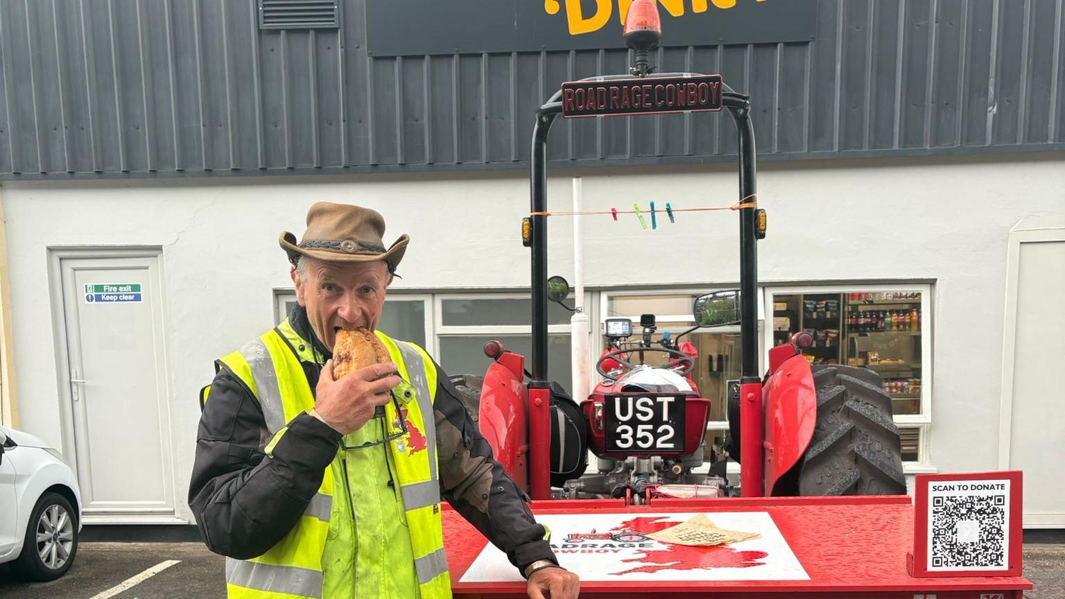 A man, wearing a cowboy hat and high visability clothing, eating a pasty. He is stood next to a red Massey Ferguson 35x tractor. The building behind them is white.