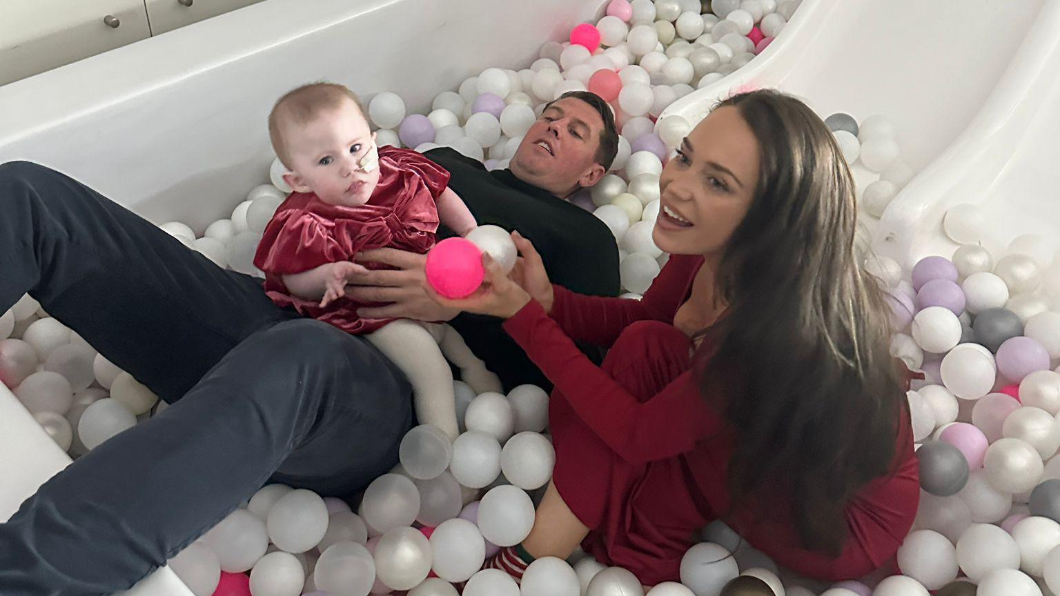 A man lying in a ball pit with a baby girl sitting on his lap and a woman sitting next to him. He is wearing a black jumper and grey trousers. The woman and baby are wearing red dresses.