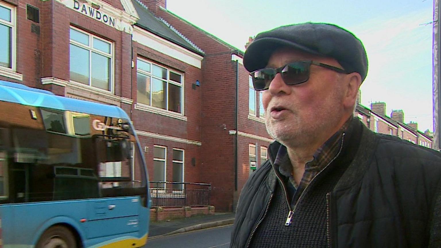 A grab taken from a TV package of Bob Arthur, an older man wearing a black flat cap, sunglasses and a black jacket. He is standing in front of the hall, a red brick building, and a bus is in the background.