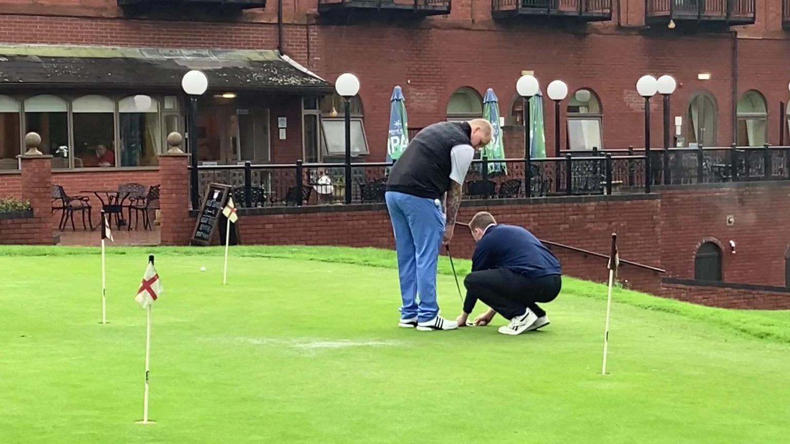 A man on a golf course holding a golf club, while another man kneels down and helps position the club