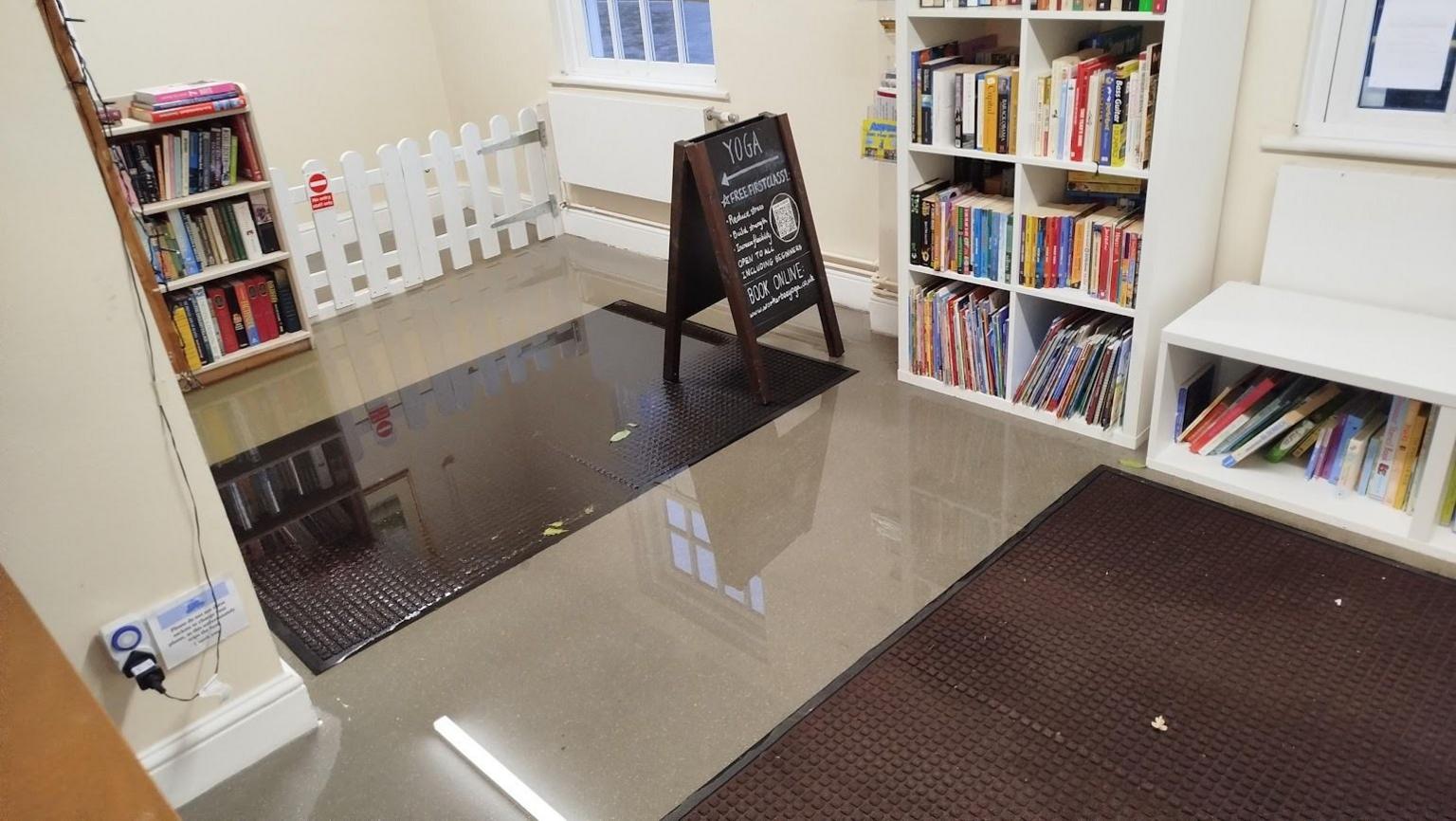 Grey and brown laminate-floored hall area with bookshelves full of books and a small A-board with writing on