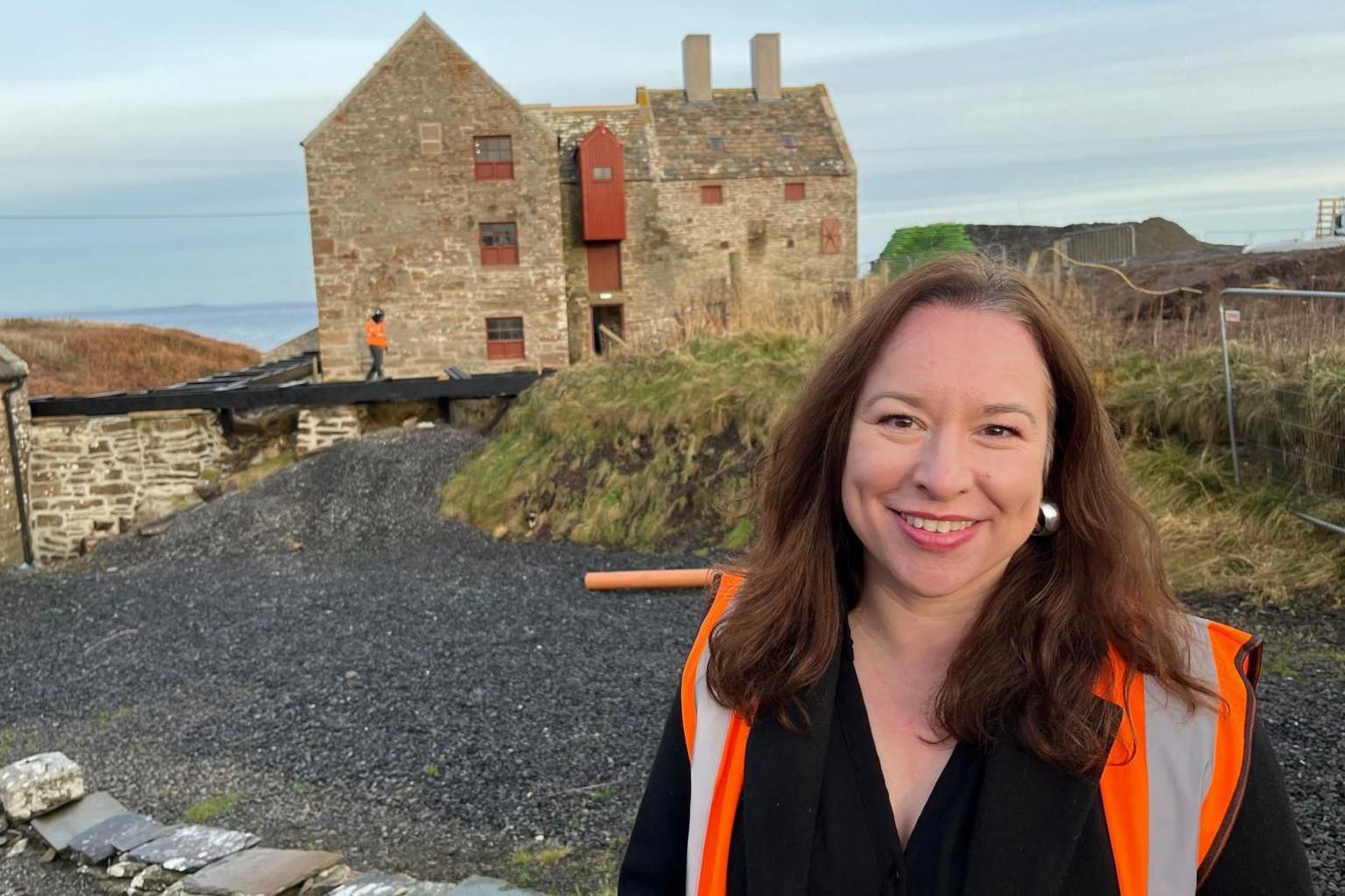 Bryony Robinson is wearing a Hi-Viz jacket and is standing in front of the historic John O' Groats mill.