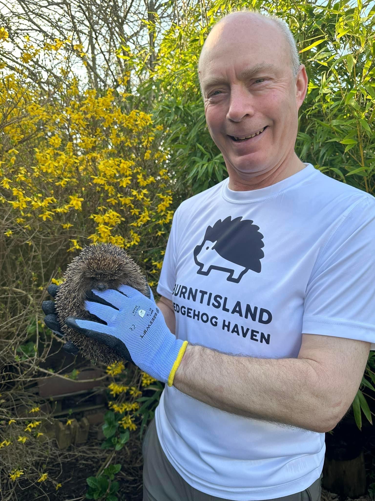 A man with light very short shaved hair wears a white tshirt with Burntisland Hedgehog Haven written on it. He is wearing blue gloves and holding a hedgehog.
