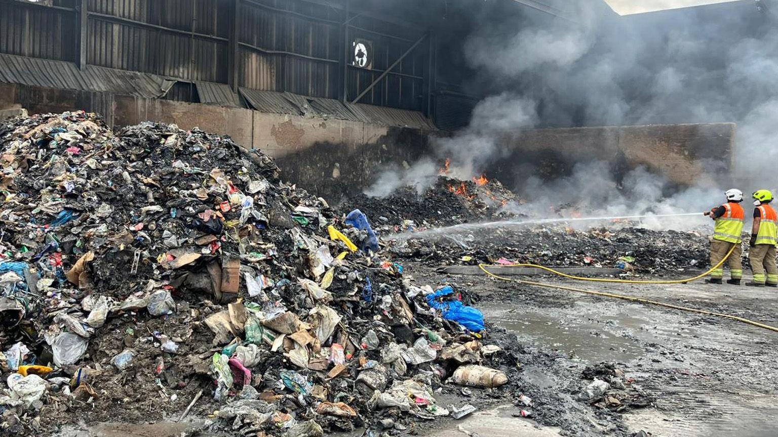 Two firefighters, holding a water hose, pointing it at rubbish and flames, they are dressed in full uniform, including helmets. Large amounts of rubbish are piled up around them.