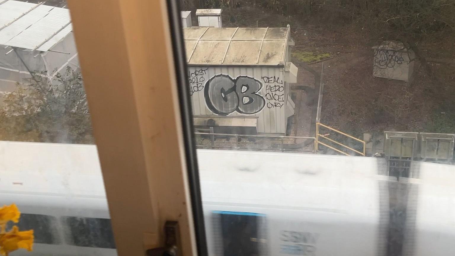 The view through a dirty window. You can see a patch of scrub land with two small dirty white buildings. There is a train going past, but it is out of focus and you can only see the roofs of the white carriages. 