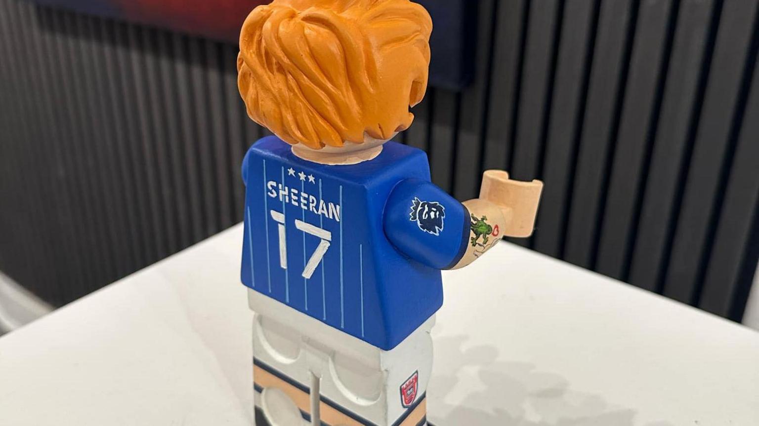 A view of the Ed Sheeran Lego figure from behind. Sheeran and the number 17 are painted on the back in white paint. The figure is sitting on a table.