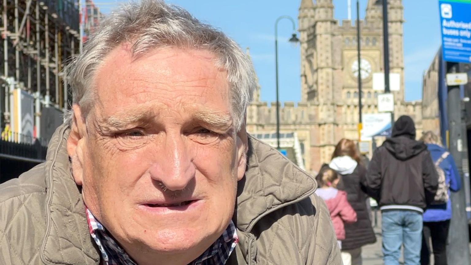Images shows an older man wearing a beige coat and checked shirt. He is looking directly at the camera. Behind him you can see people walking by. It looks like he is in a city centre location with a large period building behind him.