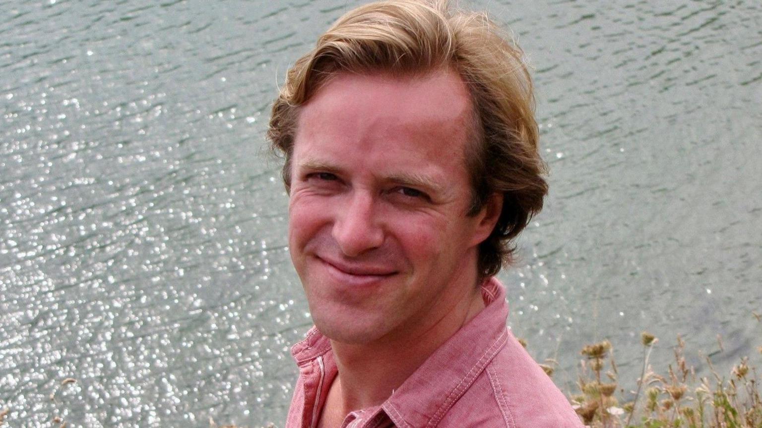 An image of Thomas Kingston, the husband of Lady Gabriella, wearing a pink shirt smiling at the camera, standing in front of the sea.