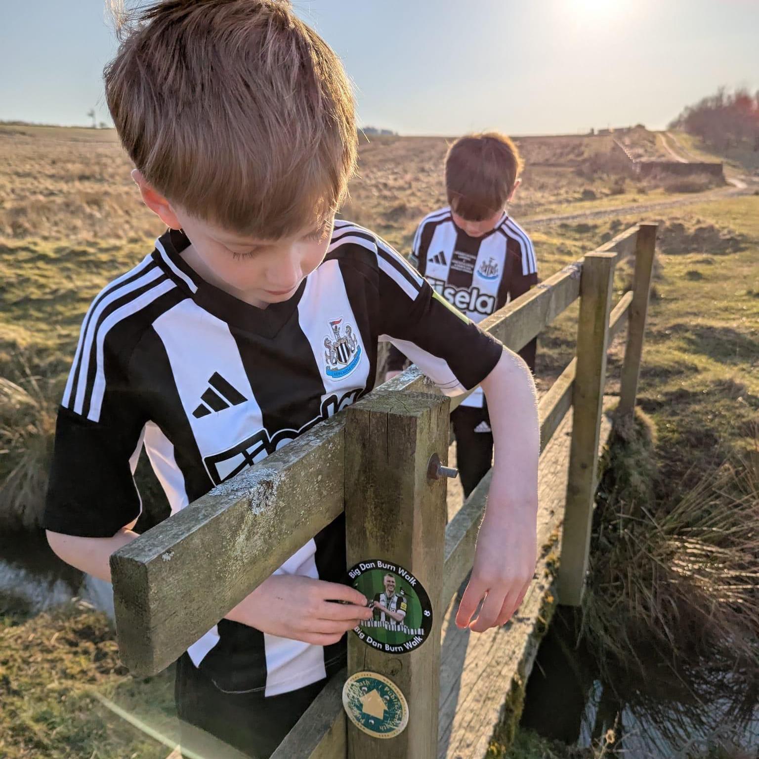 Two young boys in their black and white Newcastle kits are looking at the 'Big Dan Burn Walk' sticker near the route marker.