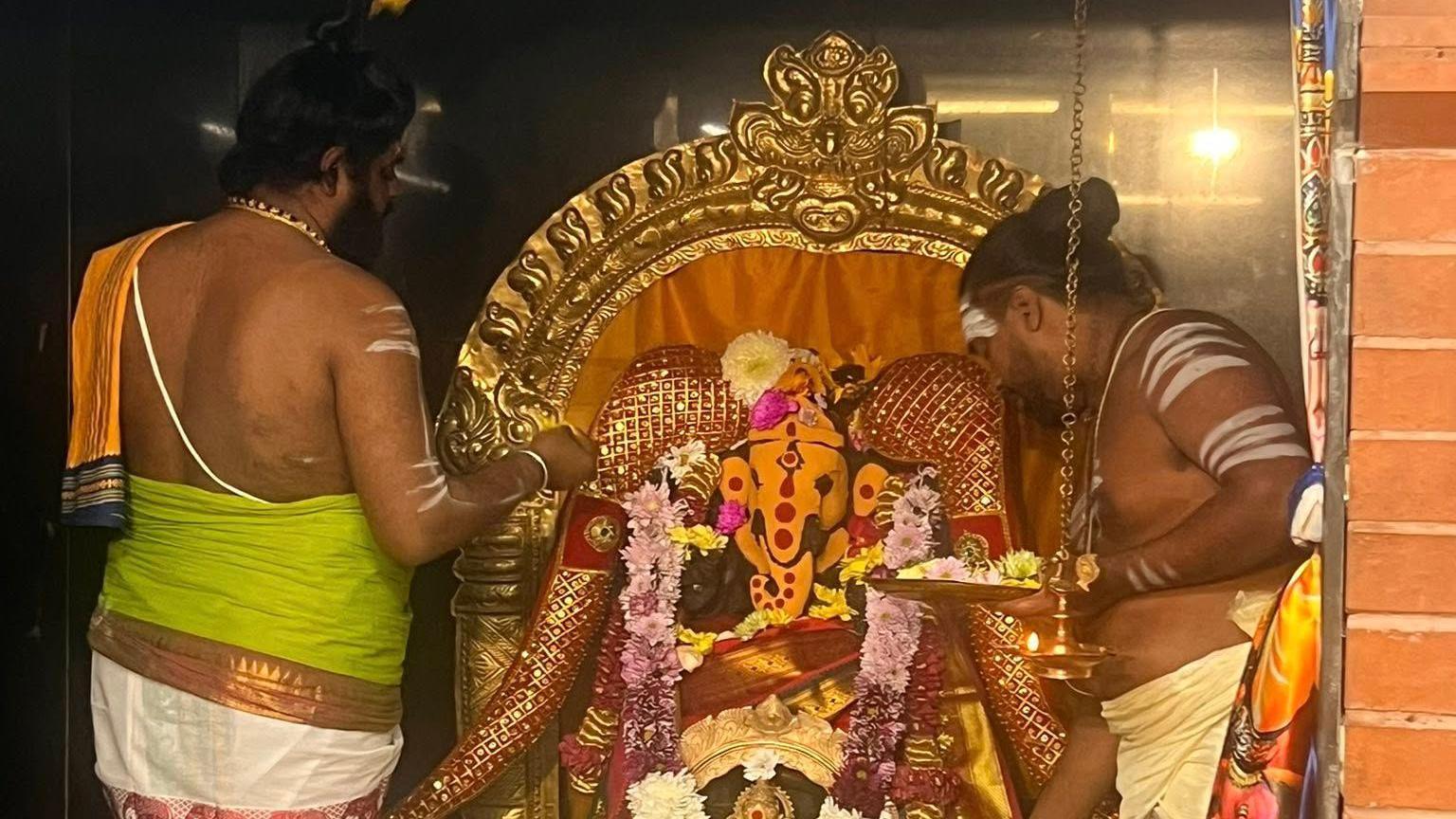 Two men dressed in traditional Indian dress with white stripes painted on their arms hold plates of flowers as part of a Hindu pooja ceremony.