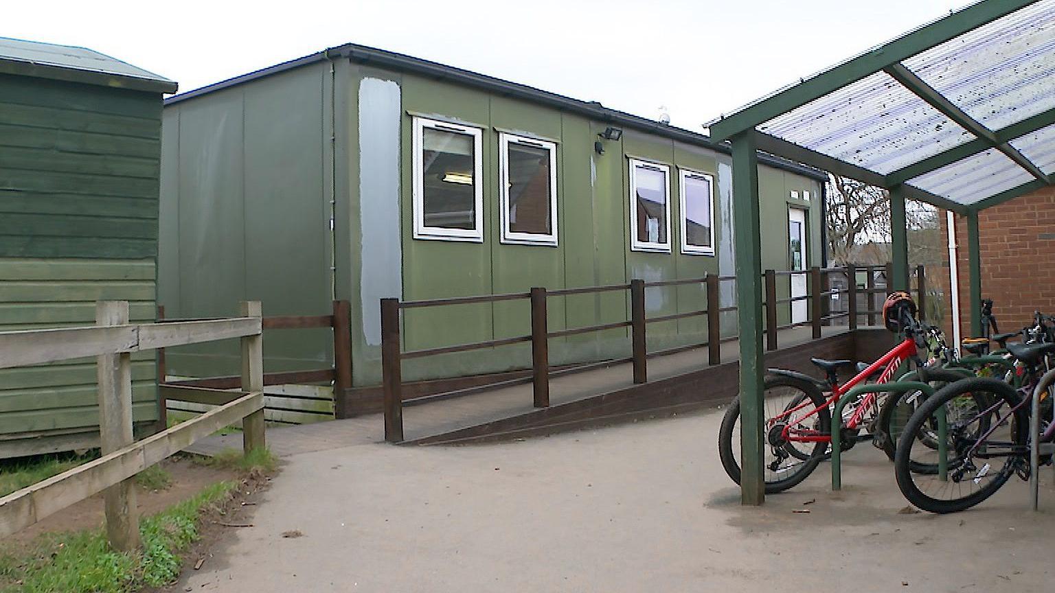 Green flat-roofed building behind a bike shed which is one of two temporary structures providing space for three classrooms