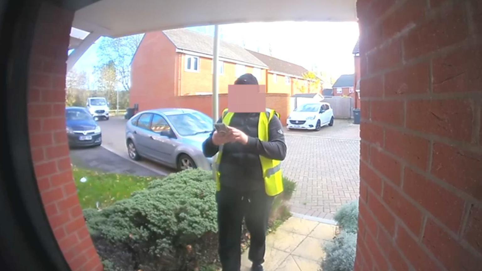 A picture taken from a doorbell camera showing a man wearing a high-viz jacket and a black hat. He is standing in front of a house taking a picture on his mobile phone.