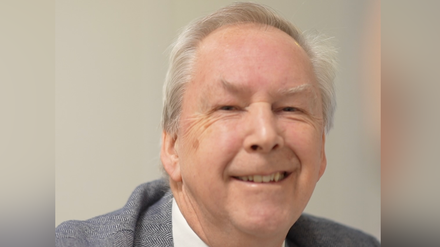 Chris Cobb smiles at the camera. He has short, thinning, grey hair. He wears a grey suit with a white shirt underneath. 