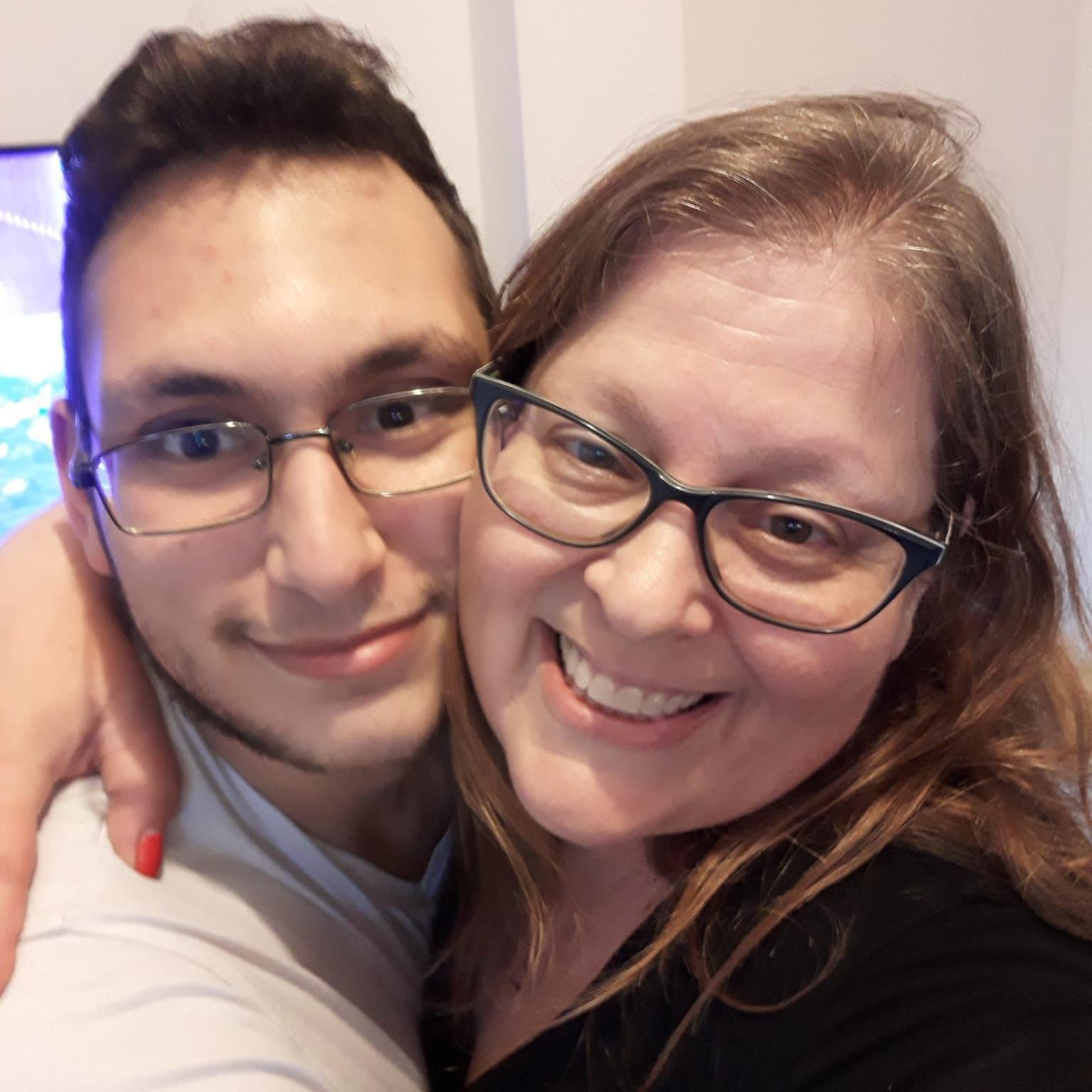 A woman and a younger man, both wearing glasses, hug and smile at the camera