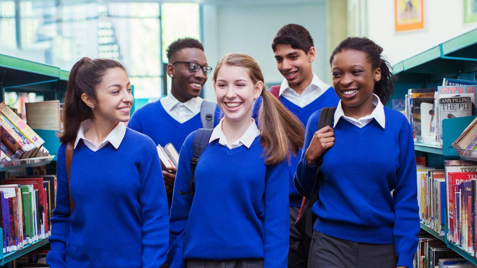 A group of five school children walk through a library while smiling wearing a white-collared shirt and a blue school jumper.