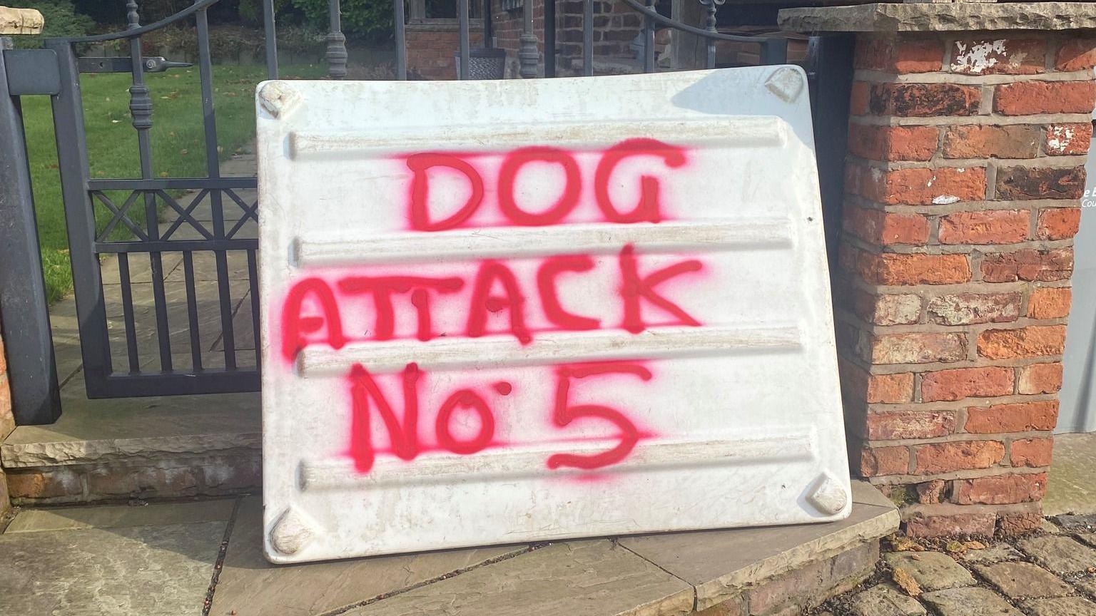 A white piece of plastic is propped up against a gate, and has been sprayed with red paint with the words 'dog attack number five'.