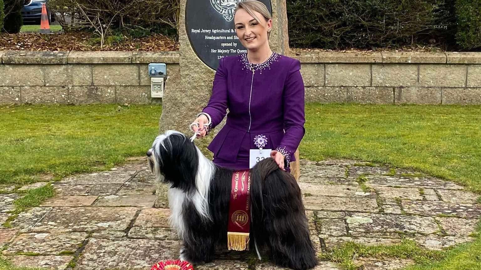 Ms Le Moignan and Mika pictured outside on some stone cobbles that are surrounded by grass. There is a stone with a plaque behind them. Mika is stood side on while wearing a red and gold sash.