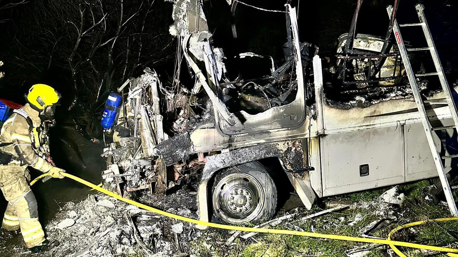 A fire fighter moving a hose reel jet around the burnt out camper van