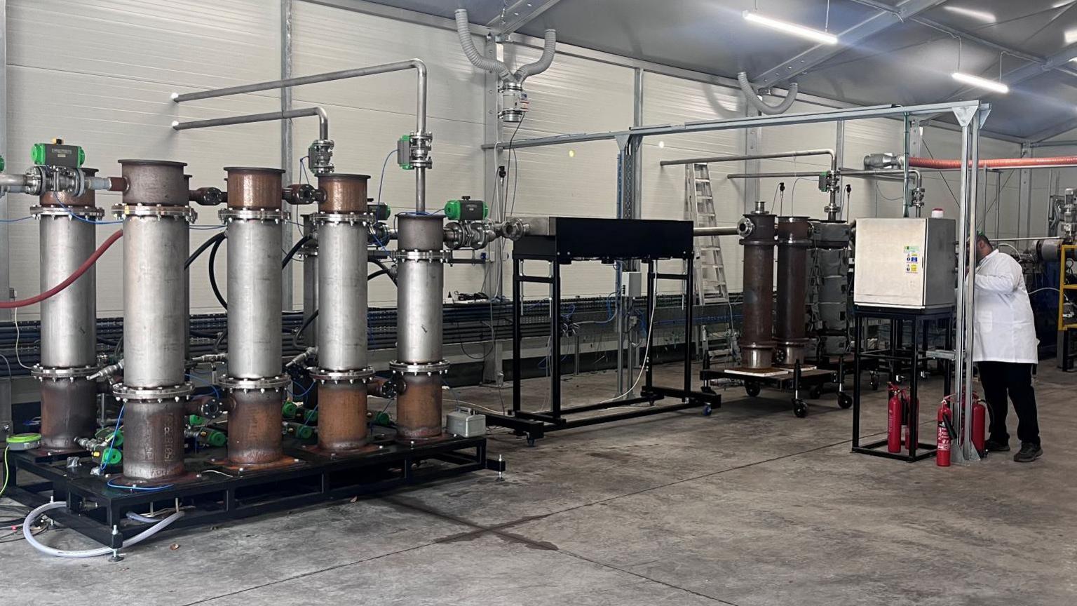 A large mechanical device with tall stacks connected to other equipment installed in a warehouse being operated by a man in a white coat