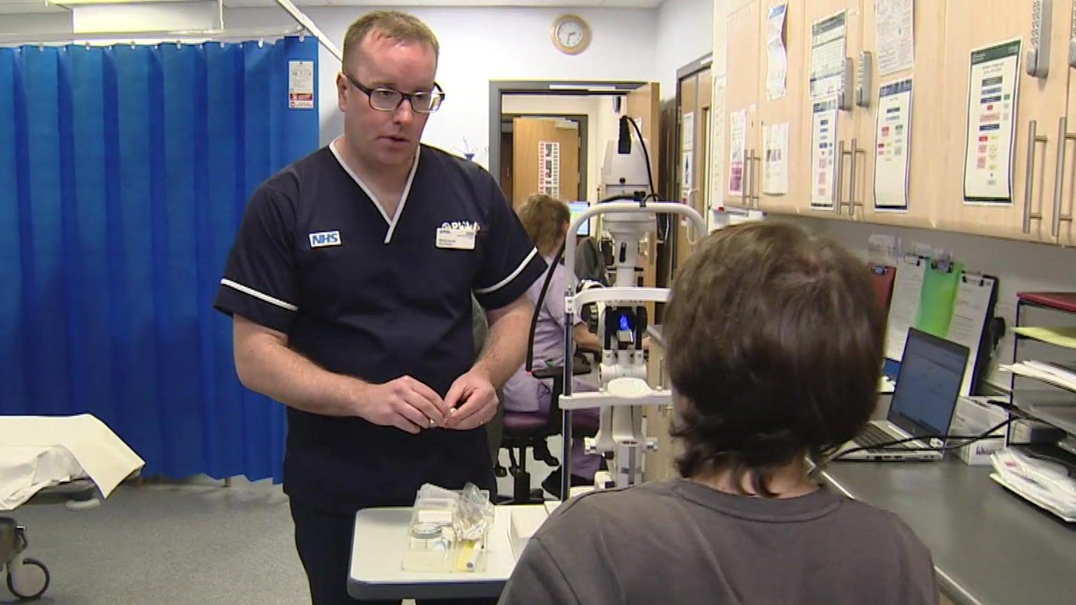 Simon Corrall, clinical manager at Lymington Urgent Treatment Centre, stands and talks to a seated patient in a screened-off treatment area. The patient had their back to the camera and Mr Corrall is facing them. He has short, brown hair, is wearing glasses and has on a navy NHS uniform with a V neck and NHS logo