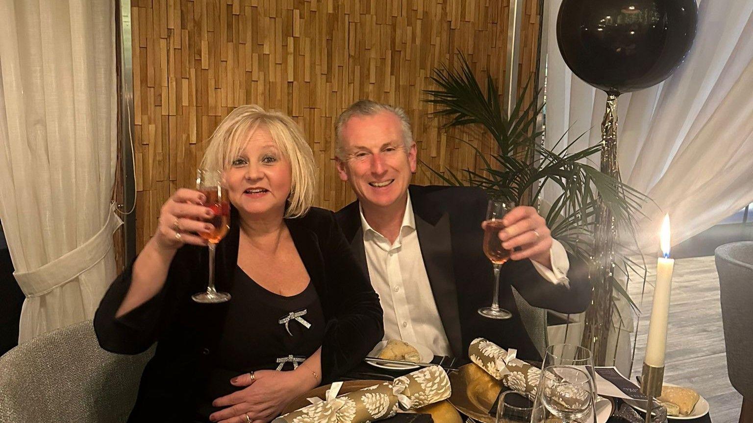 Stuart Emerson and his wife Claudia celebrating Christmas in 2024. They are sitting at a table and holding their glasses of pink champagne in the air. He is wearing a white shirt and a black blazer, while she is dressed in a black dress with silver bows and a black jacket. On the table there are gold crackers laying on the plates, and the room is decorated with candles and black and gold balloons.