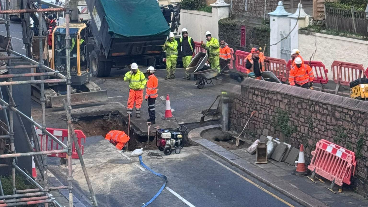 A sinkhole in a road, surrounded by equipment and workers wearing high-visibility clothing. There is scaffolding on buildings on either side of the road affected by the sinkhole, and a work truck in the vicinity.