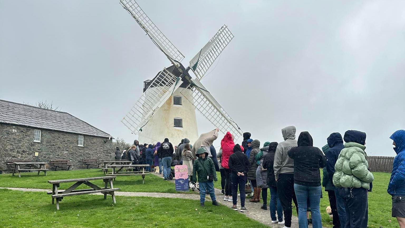A line of people queuing up in front of a white windmill. They are stood beside wooden benches.
