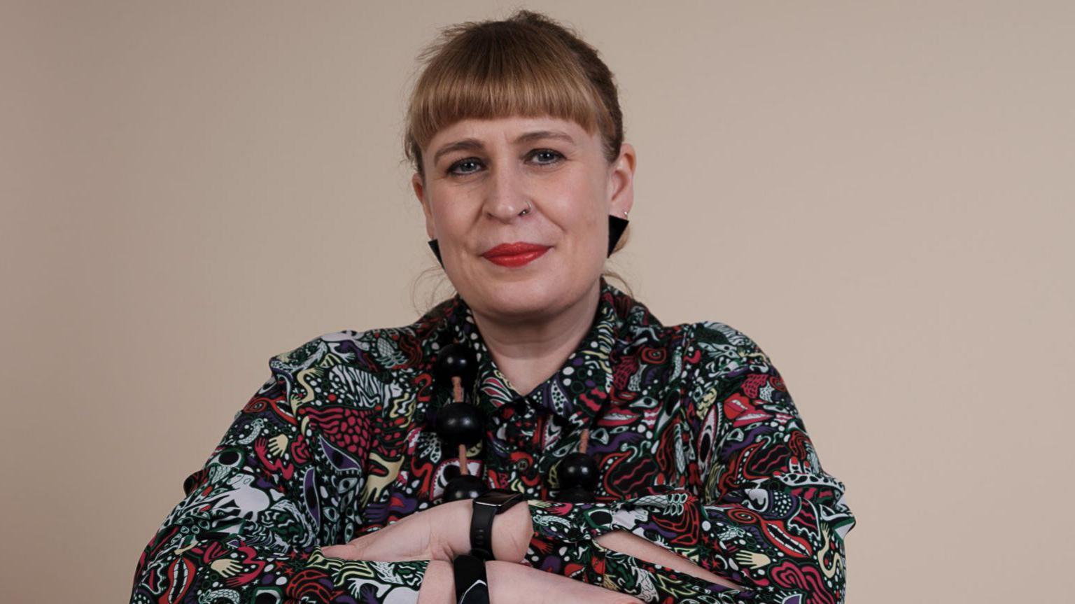A woman with blonde hair tied back. She is wearing a multi-coloured patterned top and has her arms folded. 