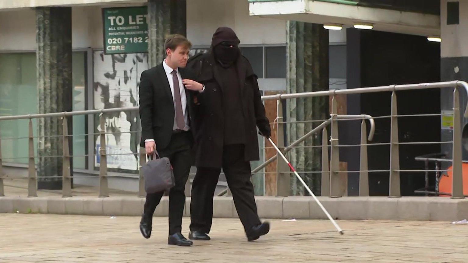 Sean Pullen, who wears a hat, a black face covering and dark glasses, walks towards the court building with a white case extended in front of him and linking arms with a younger man in a black suit carrying a laptop bag