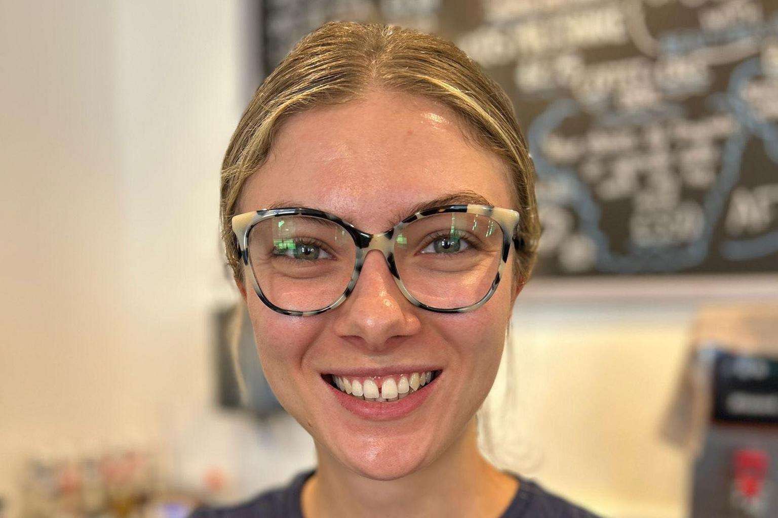 A woman with blonde-hair pulled back and large glasses wearing a dark blue round-necked T-shirt