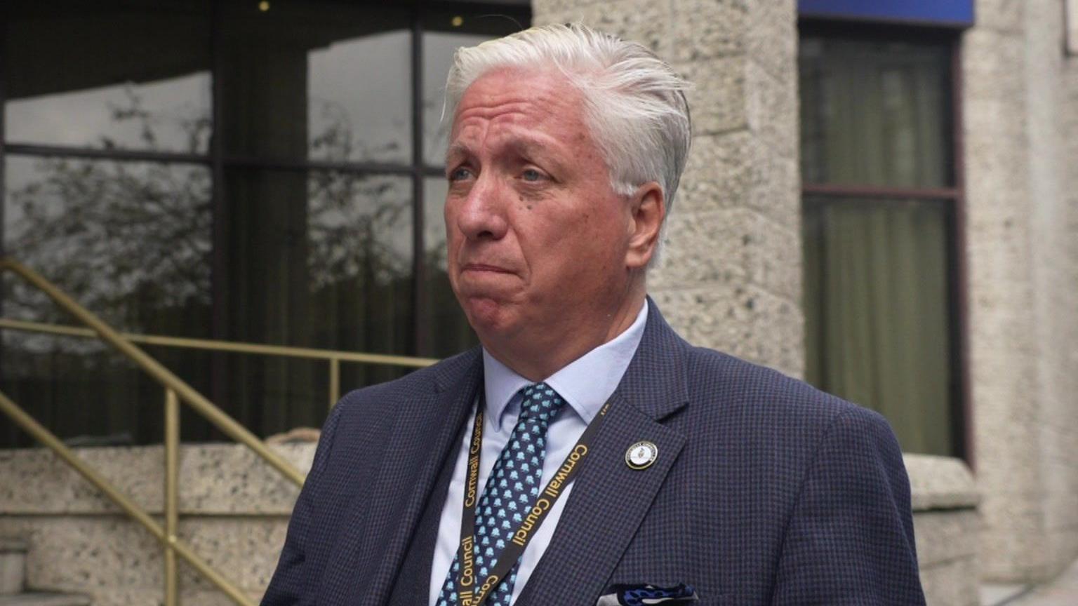 Martin Worth wears a navy blue suit and tie with light blue squares on it. He has a lanyard with Cornwall Council written on it and is standing in front of a building.