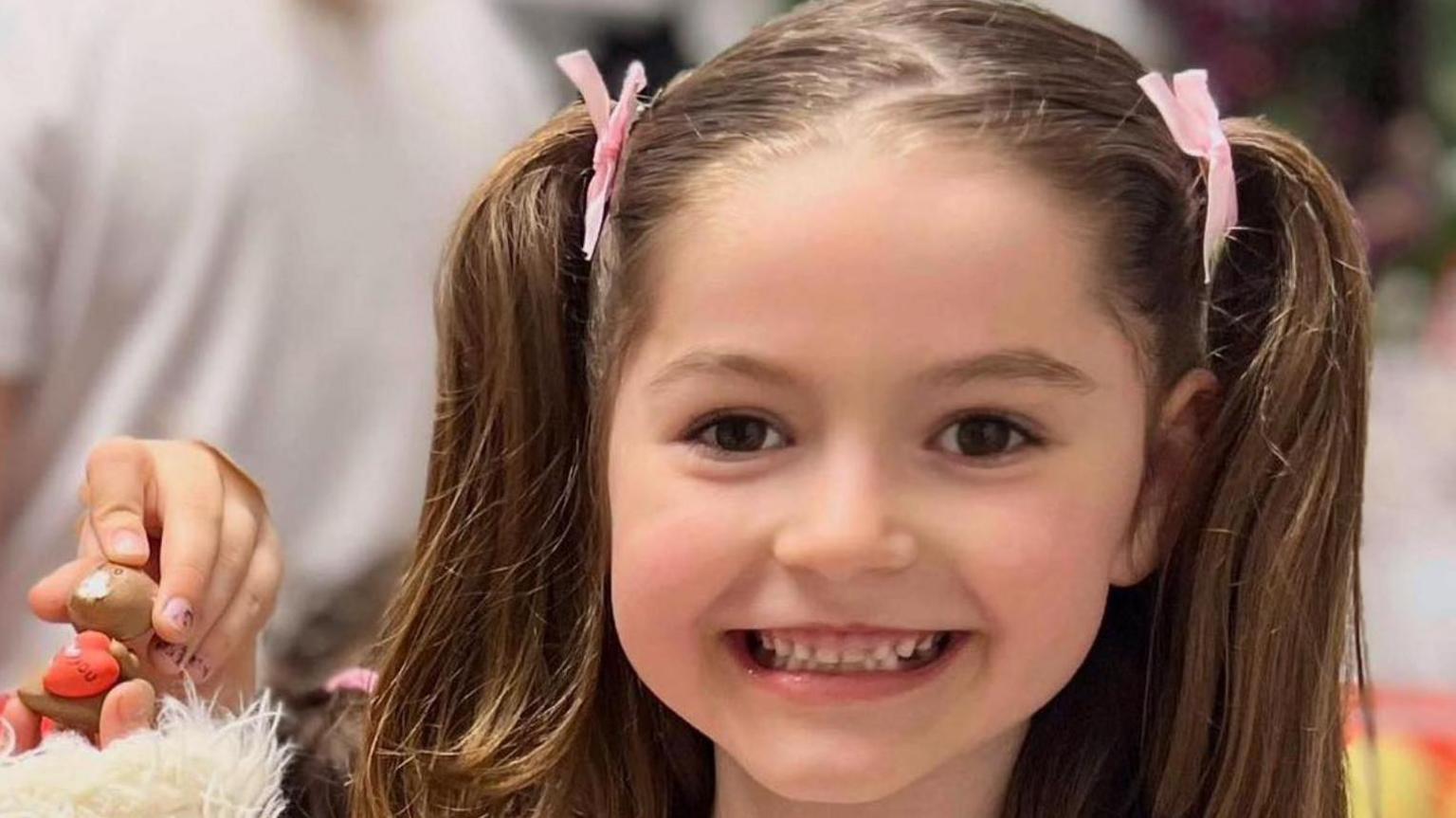 A little girl beaming at the camera. She is wearing her hair in pigtails with pink bows to tie them.
