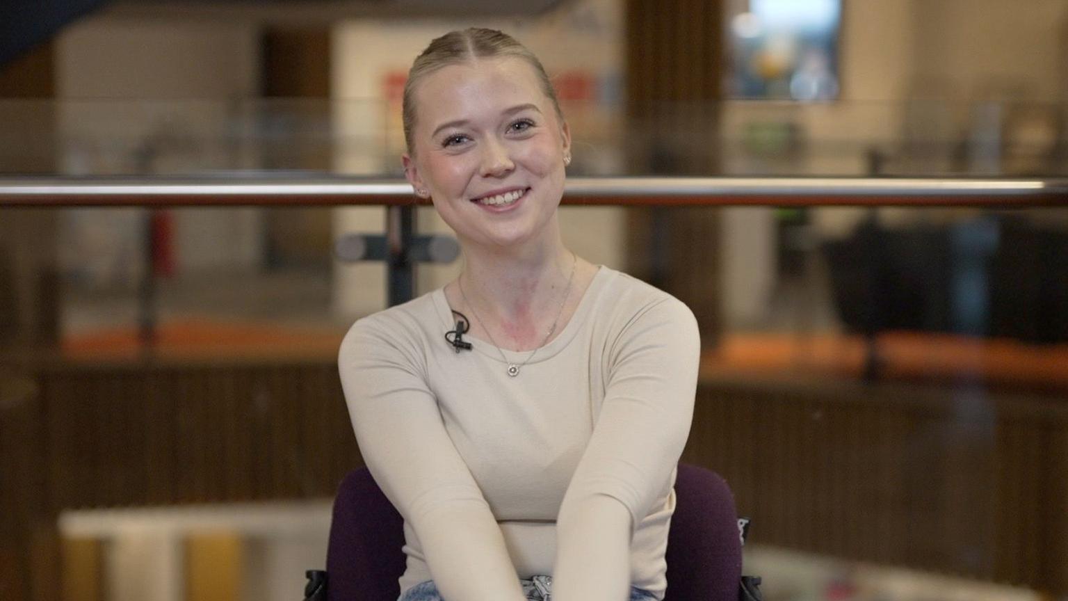 Greta Gustafsson sat at Bournemouth University. She is sitting on a purple chair wearing a long-sleeve beige top. She is smiling at the camera and has a metal necklace. The background room is blurry, but there is a metal rail running through the frame.