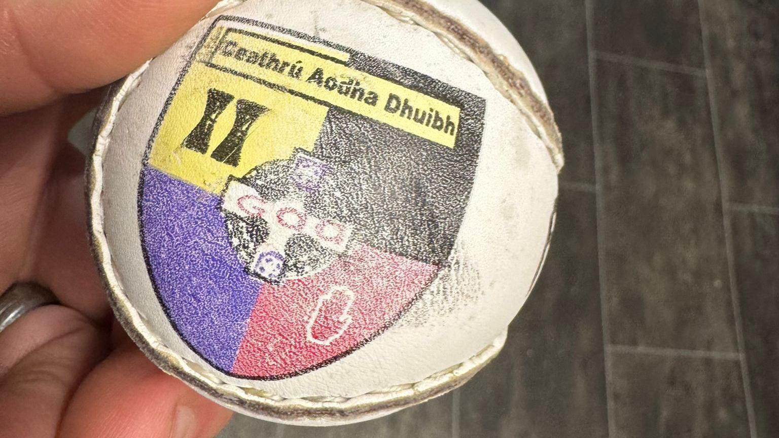 A close-up of a man's hand holding a sliotar or hurling ball embellished with the black, yellow, purple and red crest of Carryduff GAC