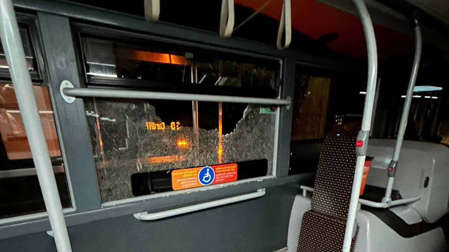 A smashed window can be seen on the bus, next to a standing area and some seats. It is dark outside.