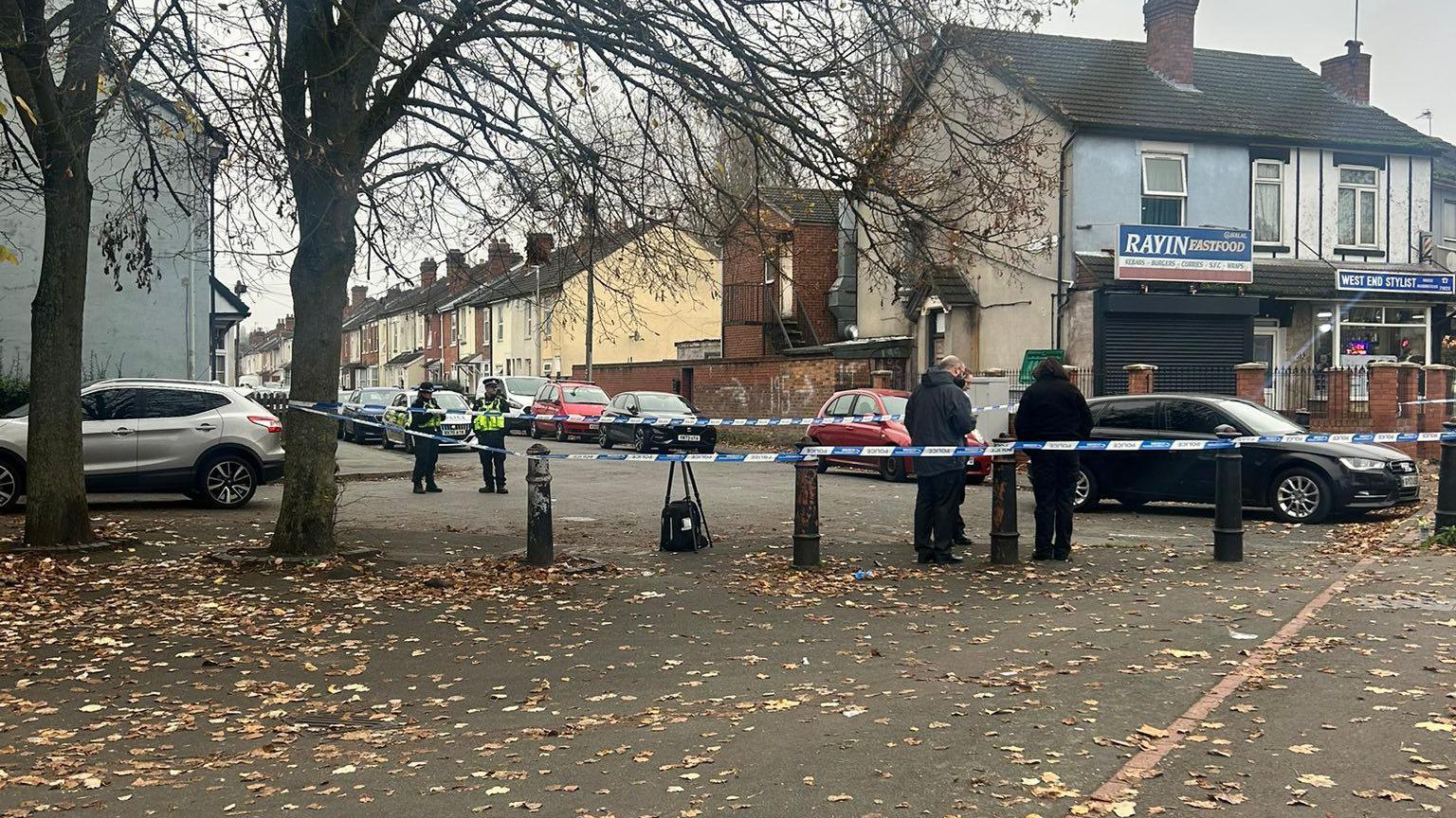 A police cordon. Officers are standing around. There is a black car near the police tape.