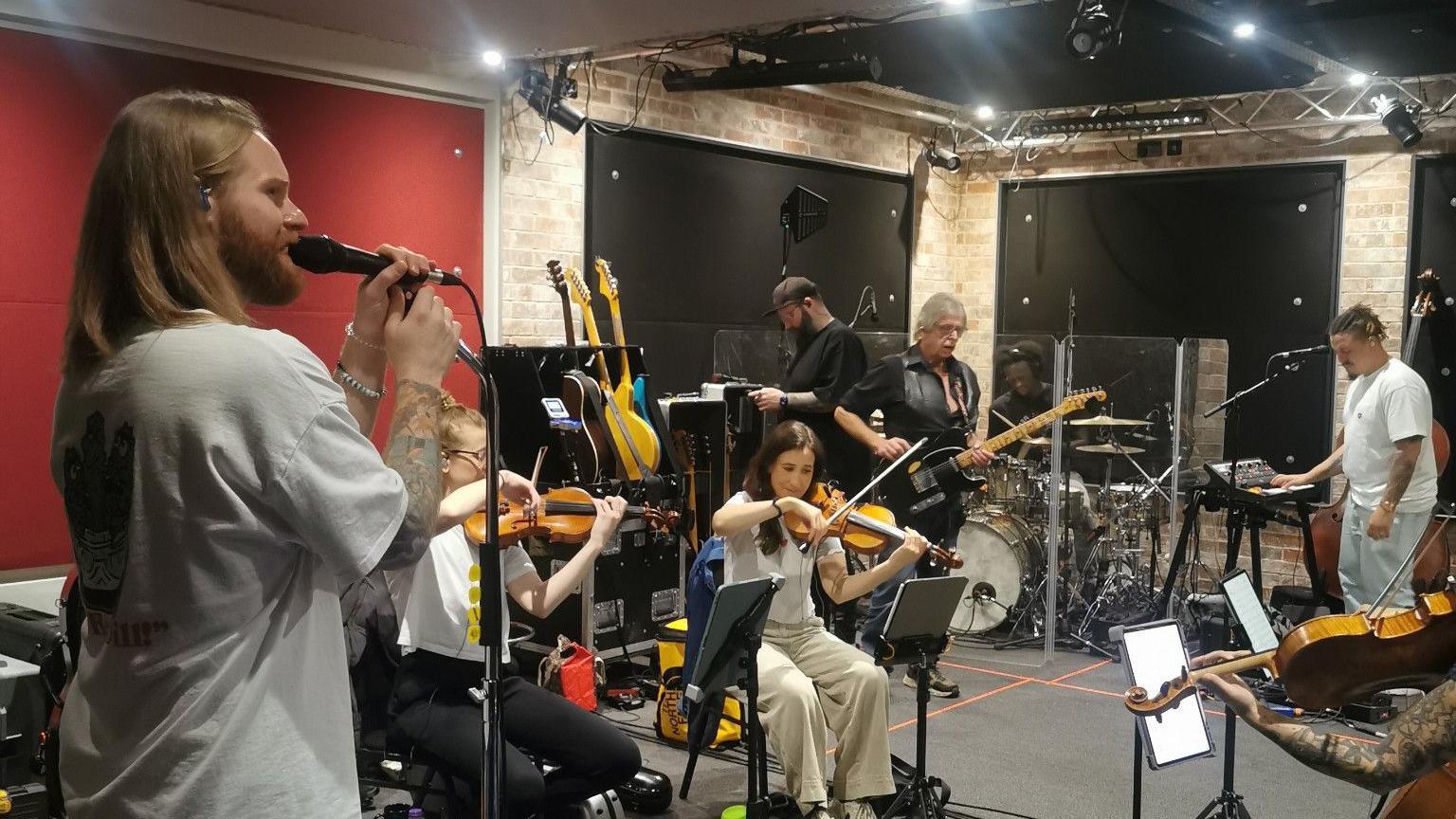 Sam Ryder singing with his live band. People are playing a violin while sitting down. While other people are playing the guitar. Sam is on the left of the picture and holding a microphone. They seem to be in a studio rehearsing.