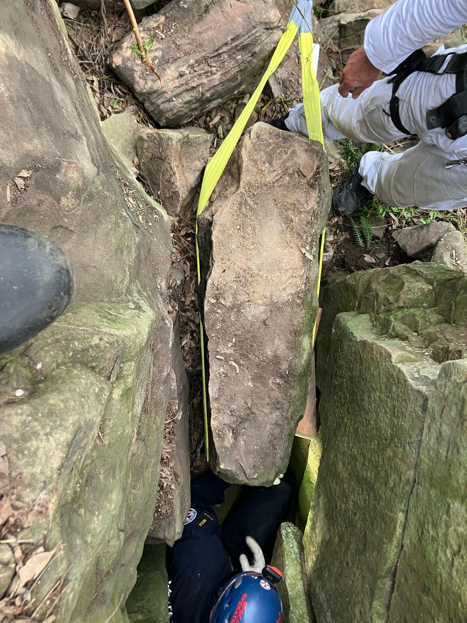 A boulder with a yellow strap around it, being lifted out from between two other boulders. A person with a helmet can be seen in the crevice, while another person's legs can be seen beside the crevice