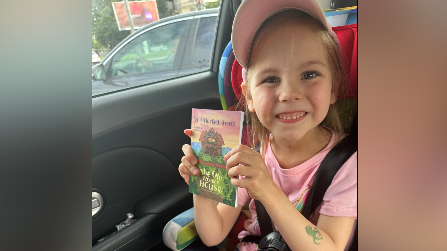 A young girl sits in a car seat smiling at the camera as she holds a book. She is wearing a pink cap and a pink T-shirt. A green fake tattoo is on her forearm in the shape of an octopus.