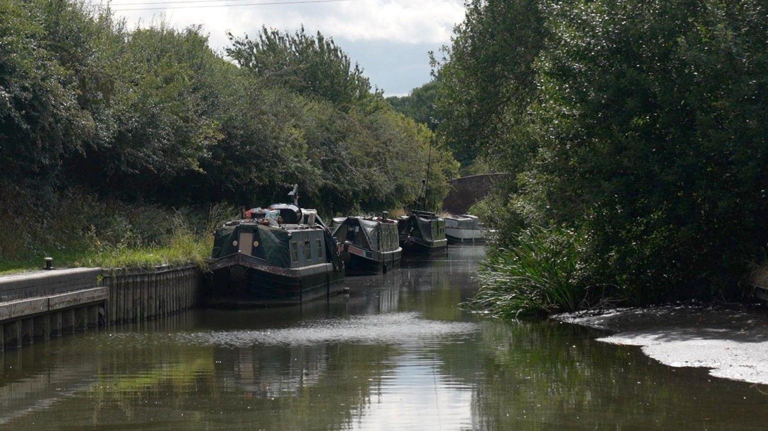 At least three narrowboats are in the background. There are trees on both sides of the canal.